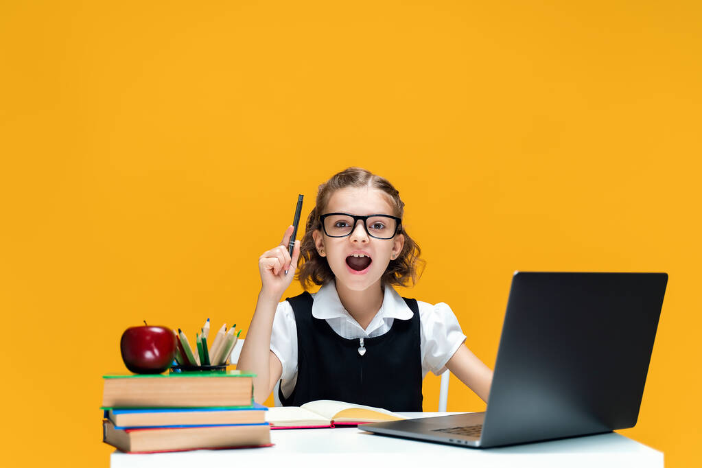 Emotioneel schoolmeisje met een bril die een pen optilt. Zittend aan het bureau met een laptop. Afstandsonderwijs  - Foto, afbeelding