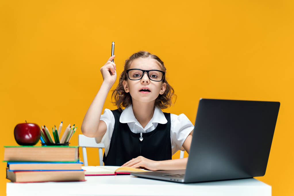 Een serieus schoolmeisje met een bril die een pen optilt. Zittend aan het bureau met een laptop. Afstandsonderwijs  - Foto, afbeelding