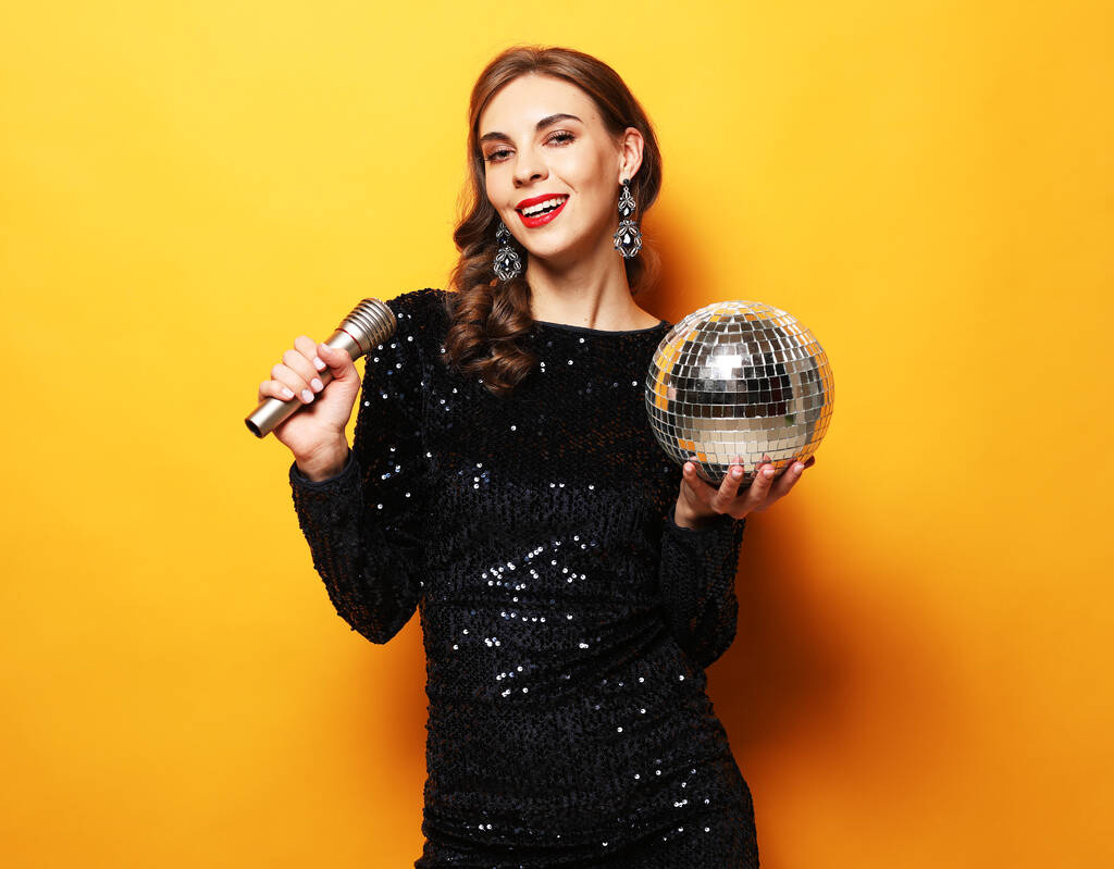 Párty, dovolená a oslavy koncepce: Mladá brunetka s dlouhými kudrnatými vlasy oblečená ve večerních šatech drží mikrofon a disco míč - Fotografie, Obrázek