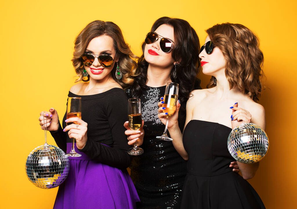 Tre gioiose affascinanti giovani donne amiche con palle da discoteca bere champagne e divertirsi insieme su sfondo giallo - Foto, immagini