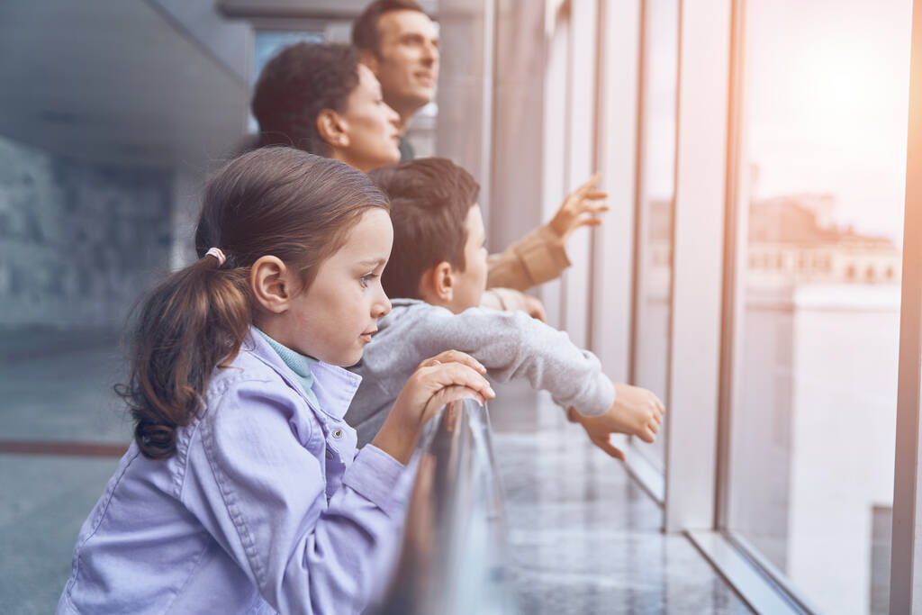Familia con dos niños pequeños mirando por una ventana mientras esperan su vuelo en la terminal del aeropuerto - Foto, imagen