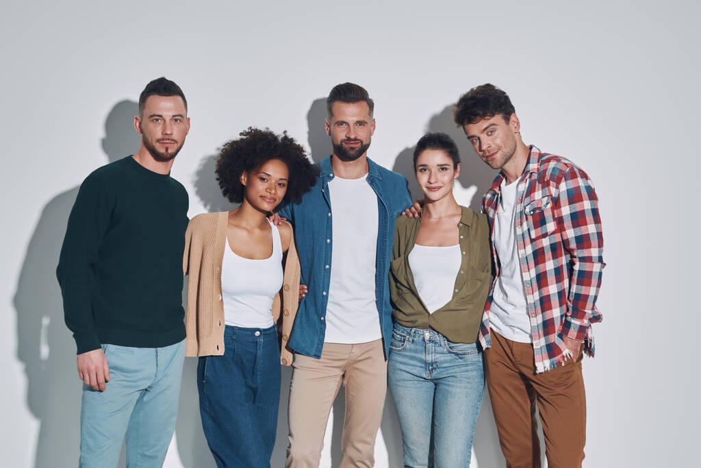 Group of young beautiful people in casual clothing bonding and smiling while standing against gray background - Photo, Image