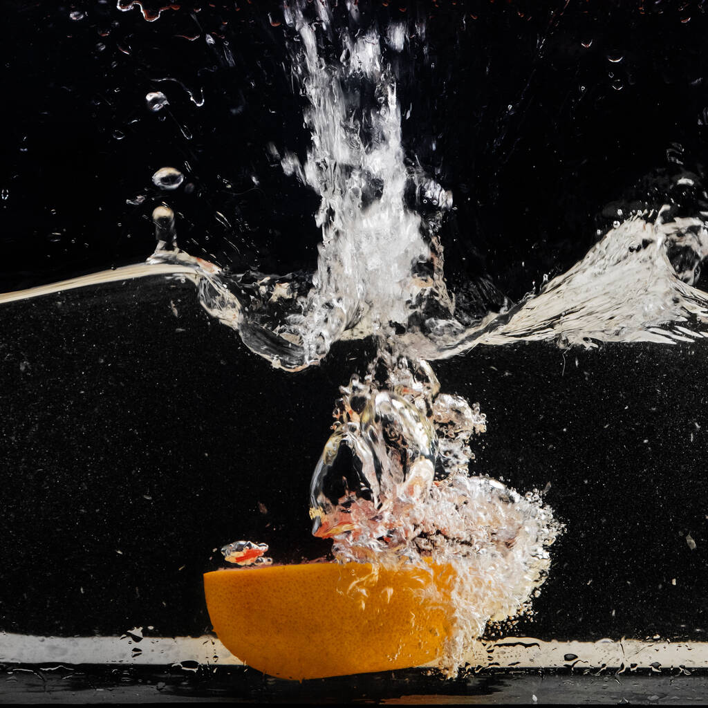 Mezza arancia fresca, fette che cadono in acqua con una spruzzata d'acqua e bolle d'aria, isolate su fondo nero. - Foto, immagini