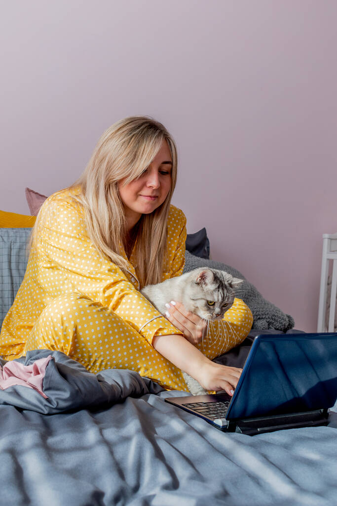 Молода жінка працює на ноутбуці, сидячи на ліжку в спальні поруч зі своїм улюбленцем. Концепція віддаленої роботи. Концепція здоров'я та психічного здоров'я
. - Фото, зображення