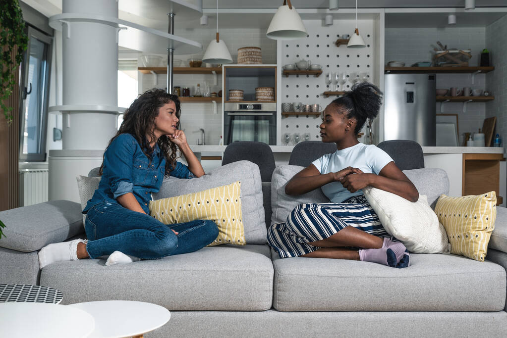 Two young white and black African roommates woman sitting on a sofa in the apartment and casually talk about rent and expenses agreement for future period before they start to work again - Photo, Image