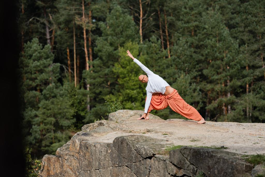 buddhist en sudadera y pantalones harem practicando yoga en postura lateral de embestida en acantilado rocoso - Foto, imagen