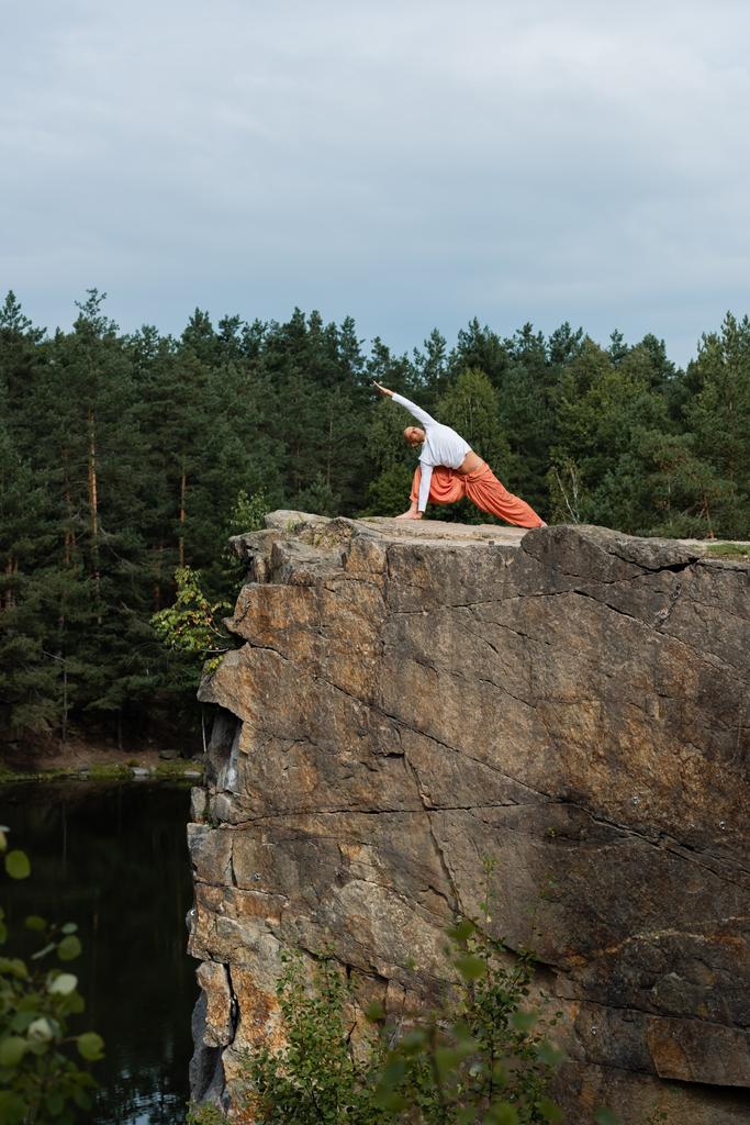 buddhist practicing side lunge pose on high rocky cliff over river - Photo, Image