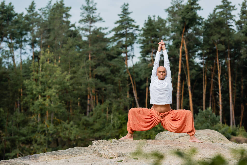 buddhist en sudadera blanca practicando pose de diosa con las manos levantadas sobre un acantilado rocoso en el bosque - Foto, imagen