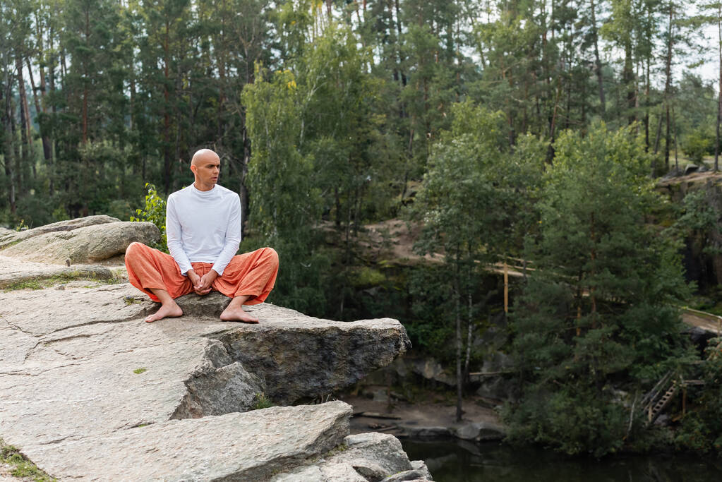 mezítlábas buddhista meditál, miközben egy sziklás sziklaszirten ül az erdei tó felett. - Fotó, kép