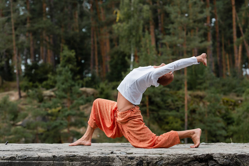 vista lateral de budista em sweatshirt e calças harém praticando crescente lunge pose ao ar livre - Foto, Imagem