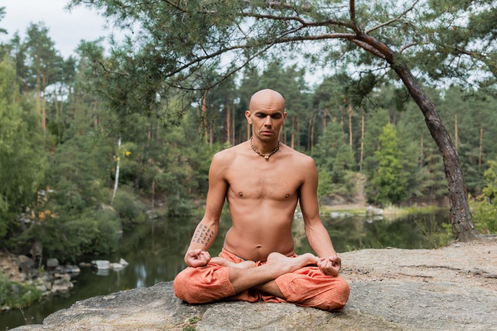 tattooed, shirtless buddhist meditating in lotus pose with closed eyes outdoors - Photo, Image
