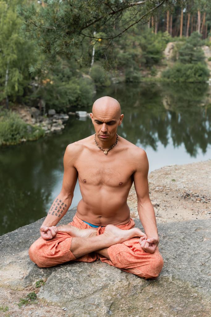 sem camisa, tatuado budista meditando em pose de lótus em penhasco rochoso sobre lago - Foto, Imagem