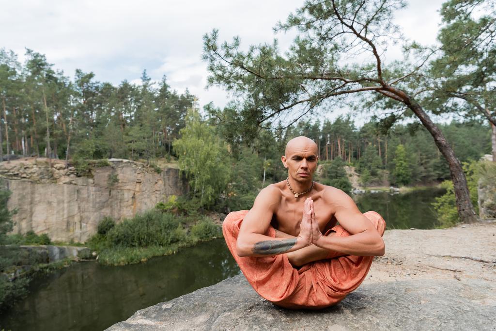 shirtless buddhist practicing extended lotus pose with praying hands on rocky cliff over river - Photo, Image