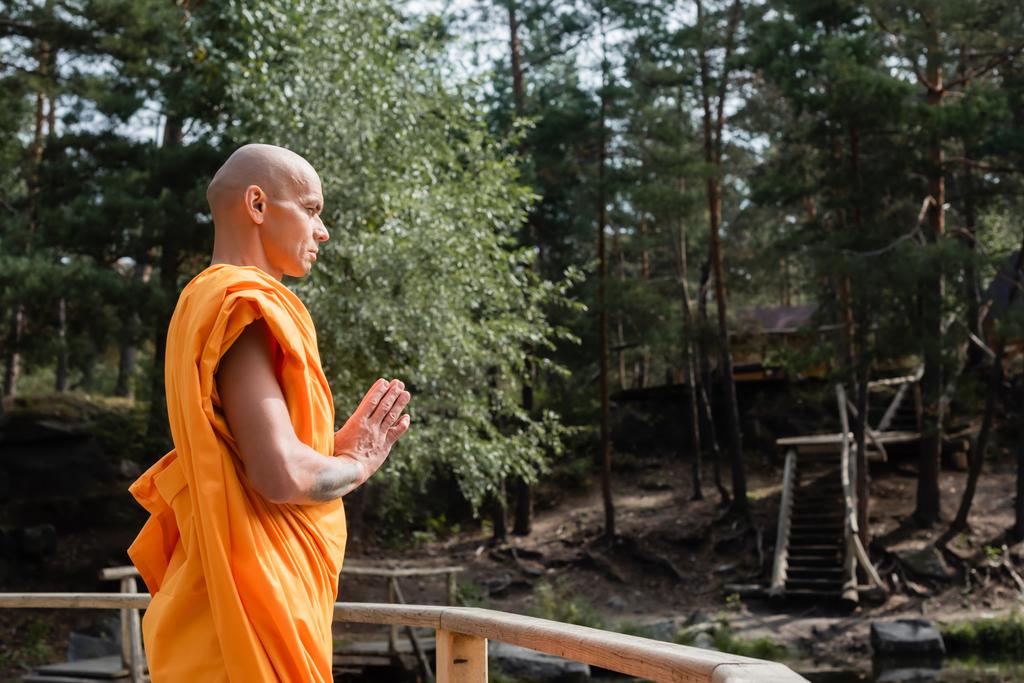 side view of buddhist meditating with praying hands near wooden fence in forest - Photo, Image