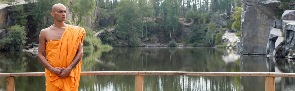 buddhist in traditional robe looking away near wooden fence over lake in forest, banner - Photo, Image
