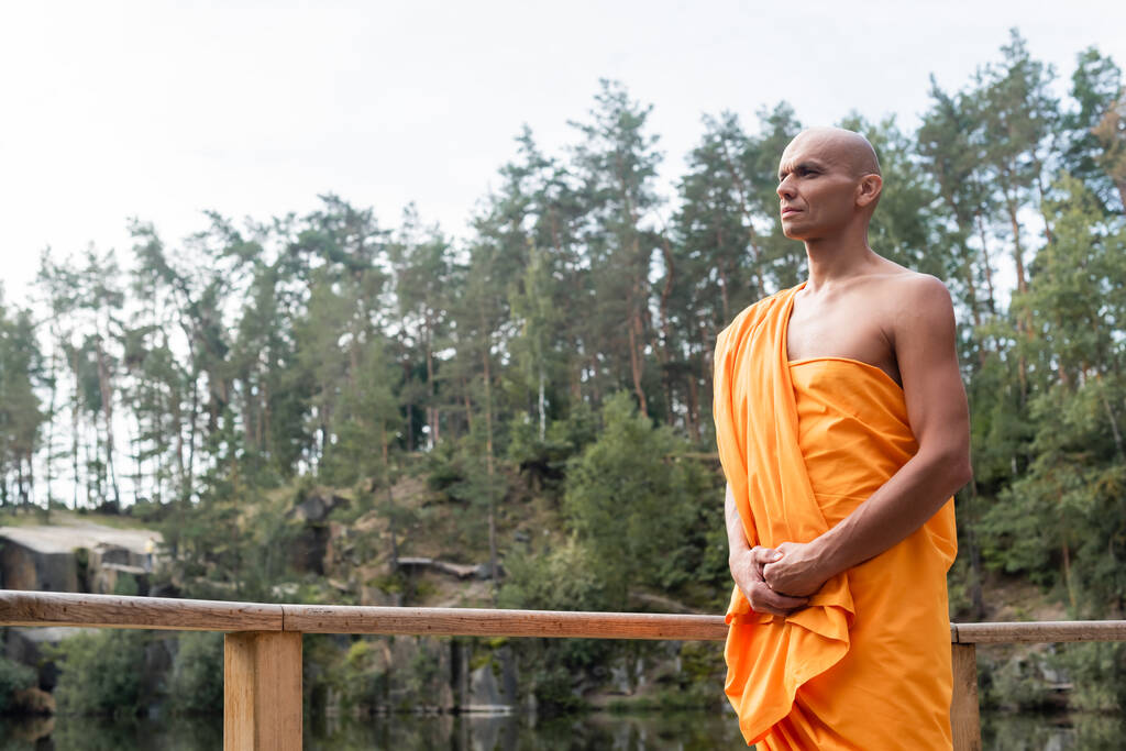 monje budista en kasaya naranja mirando hacia otro lado mientras medita cerca de una cerca de madera en el bosque - Foto, Imagen
