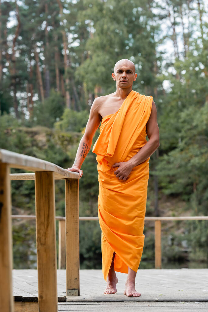 pełny widok buddyjskiego mnicha stojącego w pobliżu drewnianego ogrodzenia w lesie - Zdjęcie, obraz