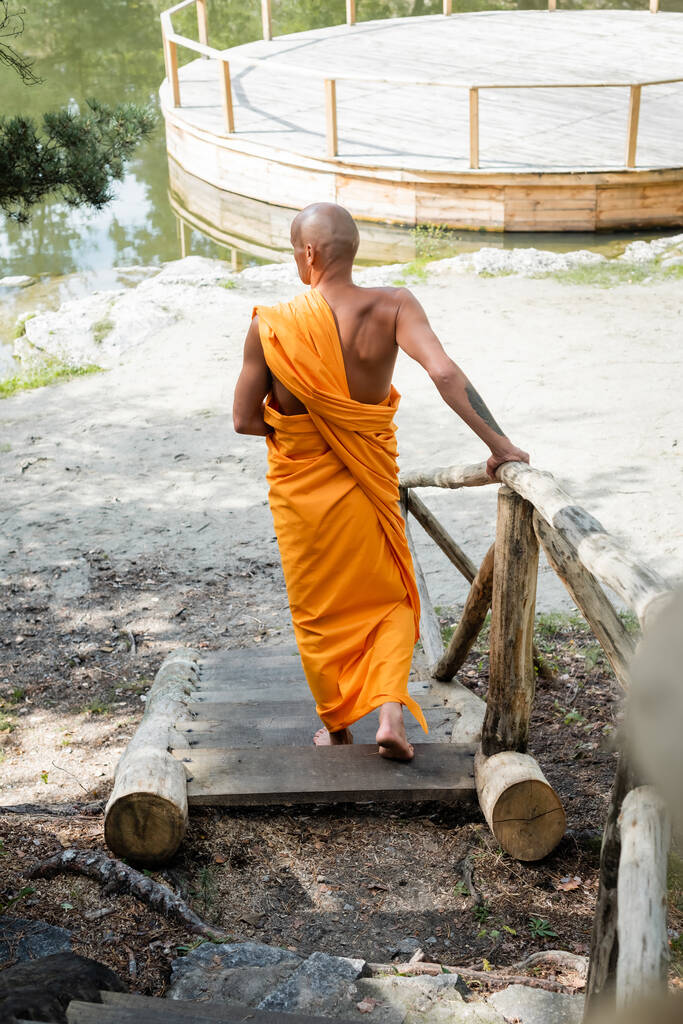 achteraanzicht van een onbehaarde man in traditionele boeddhistische kleding lopend op houten trappen in het bos - Foto, afbeelding