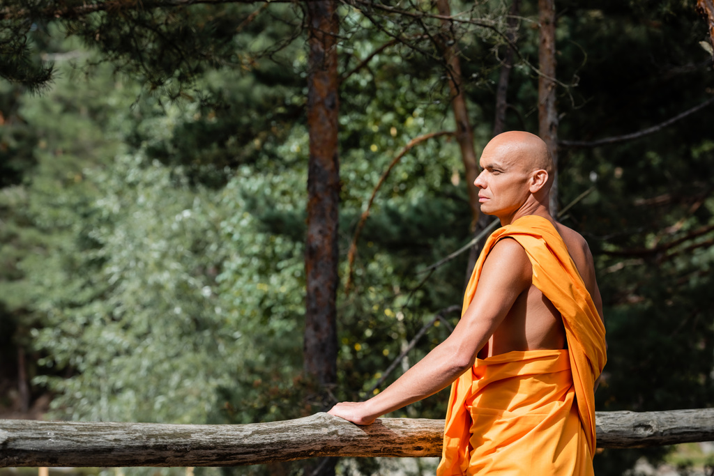 uomo in abiti tradizionali buddisti in piedi vicino alla recinzione di legno nella foresta e guardando altrove - Foto, immagini