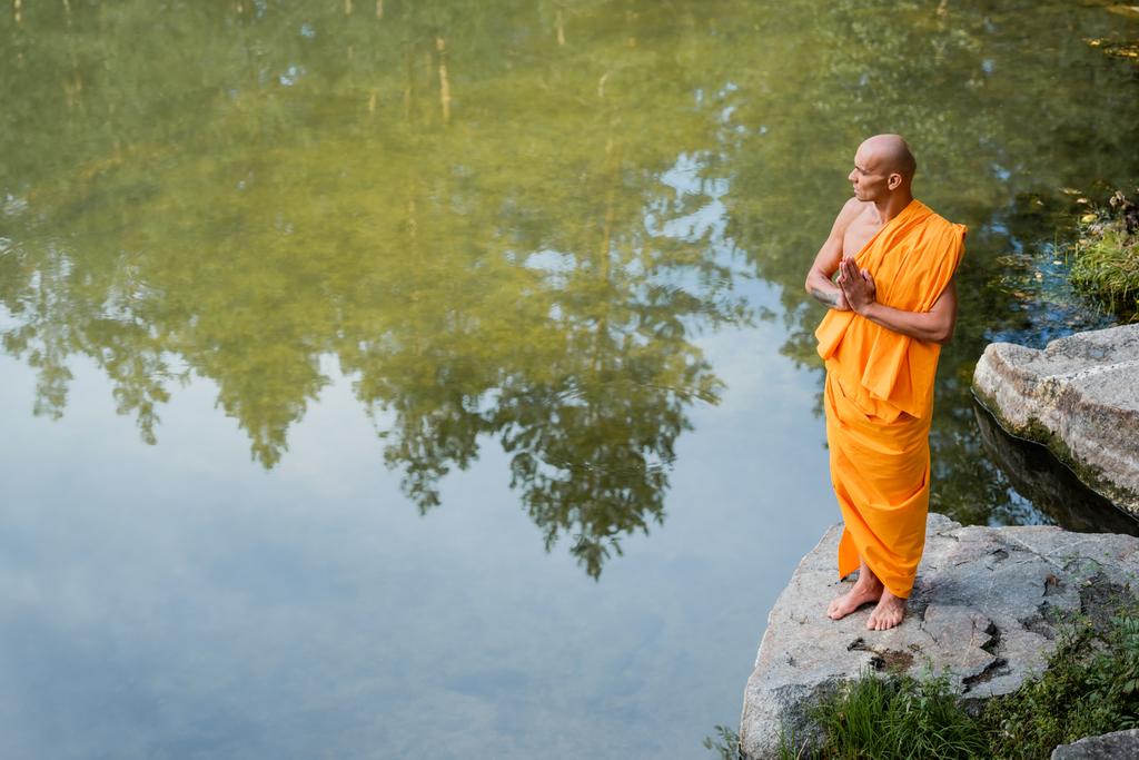 wysoki kąt widzenia buddysty w pomarańczowym kasaja medytacji z modlących się rąk w pobliżu jeziora - Zdjęcie, obraz