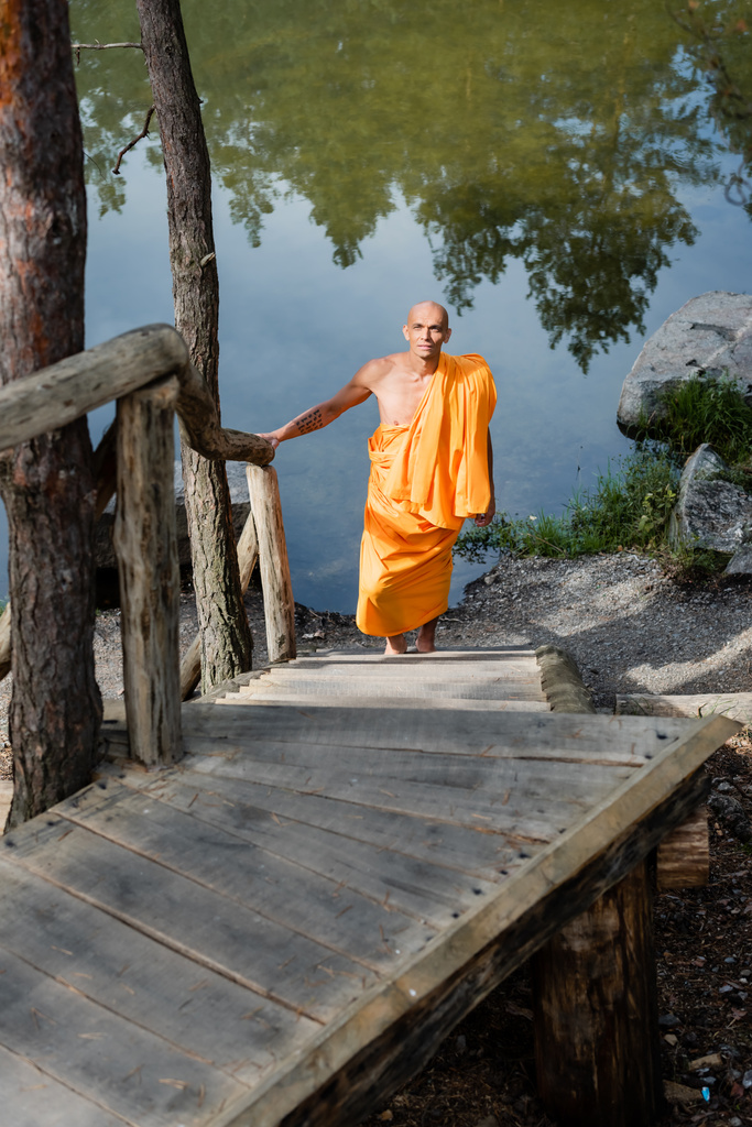 vista ad alto angolo di buddista in vestaglia arancione passeggiando su scale di legno vicino al lago - Foto, immagini