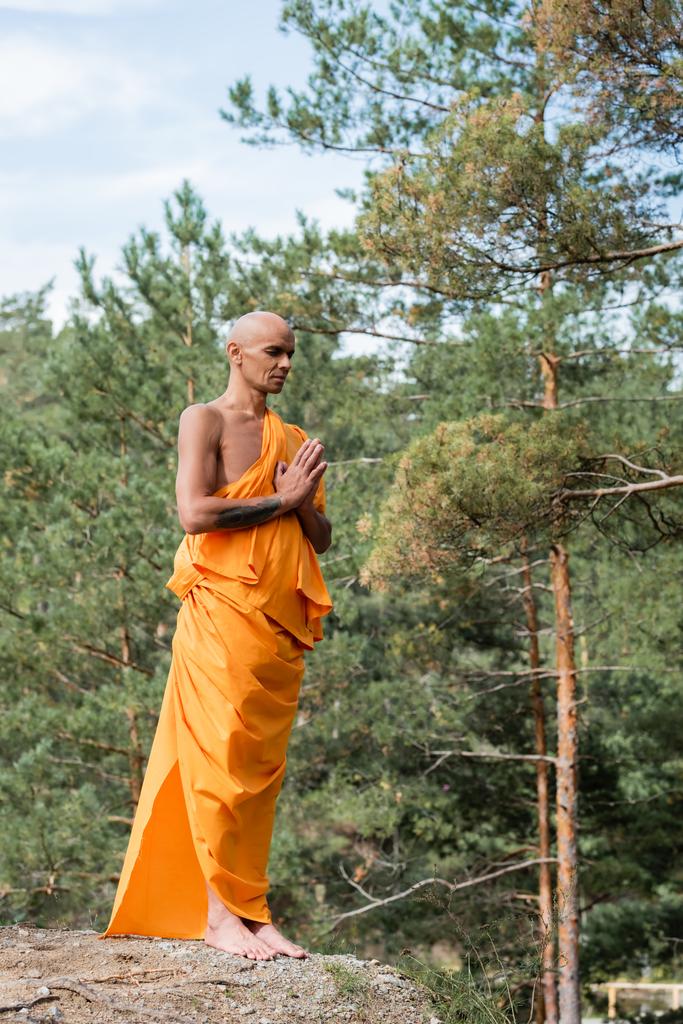 full length view of buddhist on orange kasaya praying with closed eyes in forest - Photo, Image