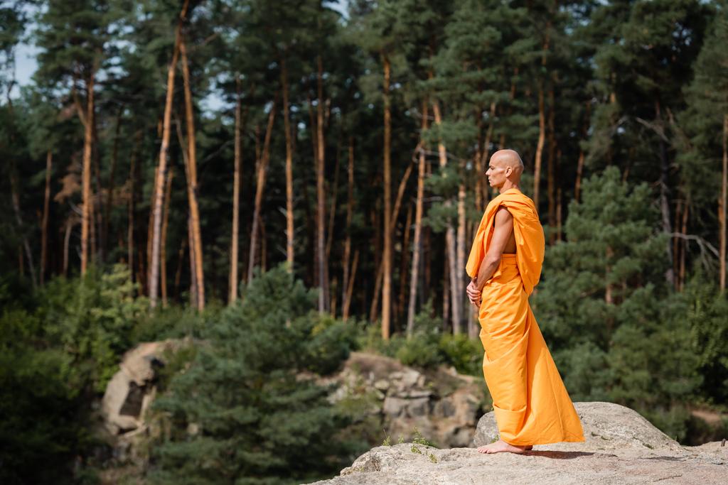 Turuncu kasaya giymiş çıplak ayaklı Budist keşişin ormandaki kayalık kayalıklarda meditasyon yapmasının yan görüntüsü. - Fotoğraf, Görsel