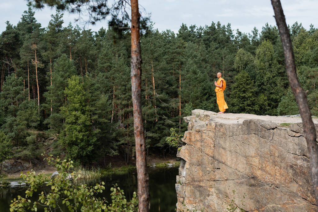 buddhalainen munkki appelsiinikasayassa rukoilemassa korkealla kalliolla joen yli metsässä - Valokuva, kuva