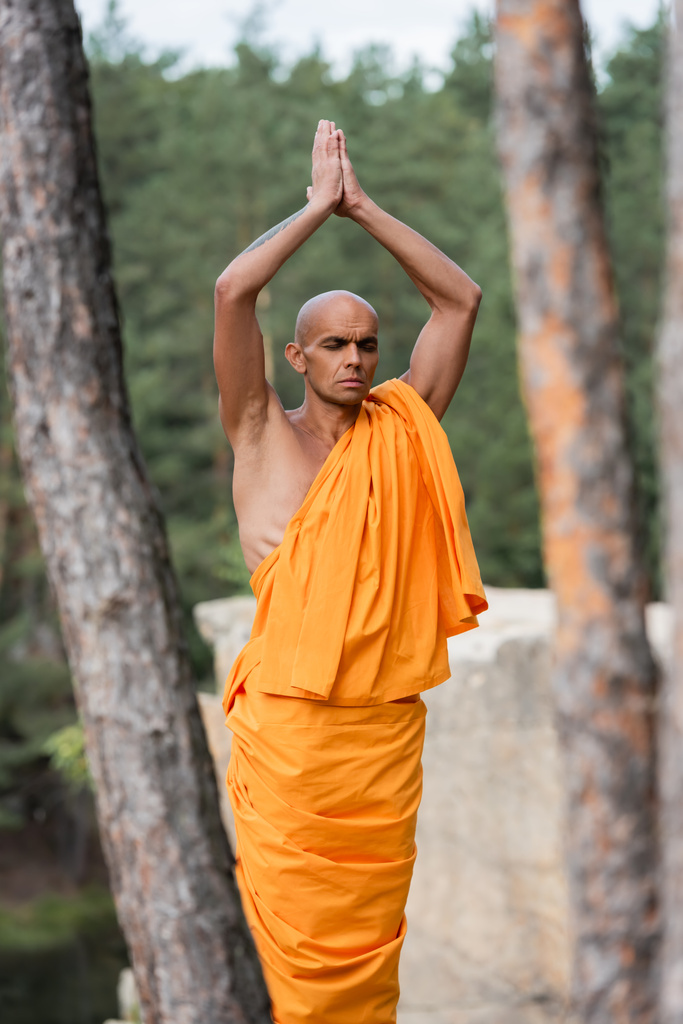 Буддийский монах с поднятыми молитвенными руками медитирует с закрытыми глазами в лесу - Фото, изображение