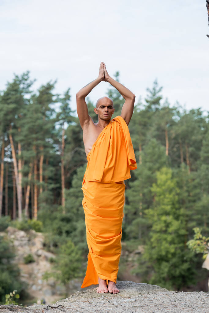 полный вид буддиста в апельсиновой касае медитирует с поднятыми молитвенными руками в лесу - Фото, изображение