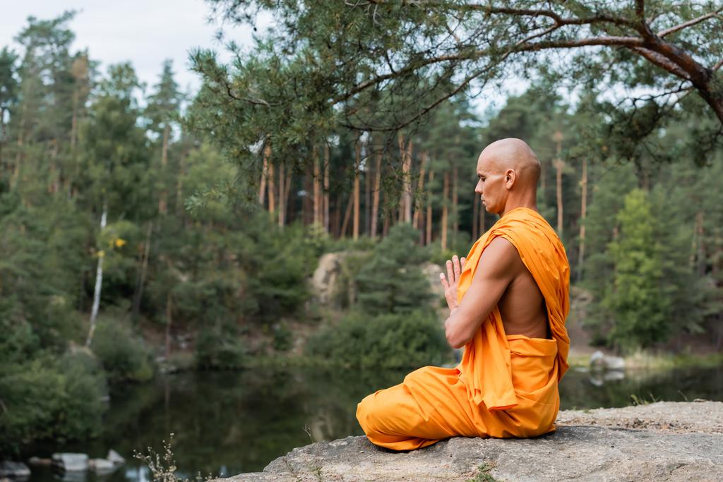 Monge budista em kasaya laranja meditando em pose de lótus com as mãos de oração perto do lago da floresta - Foto, Imagem