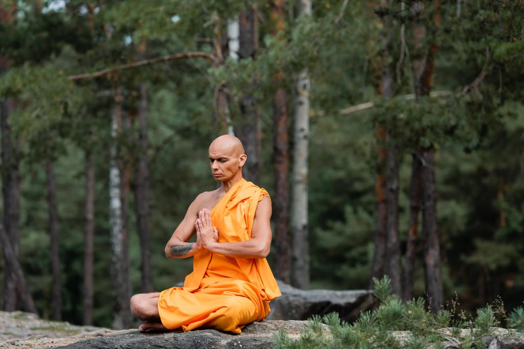 Буддийский монах в оранжевой касайе сидит в позе лотоса с молитвенными руками во время медитации в лесу - Фото, изображение