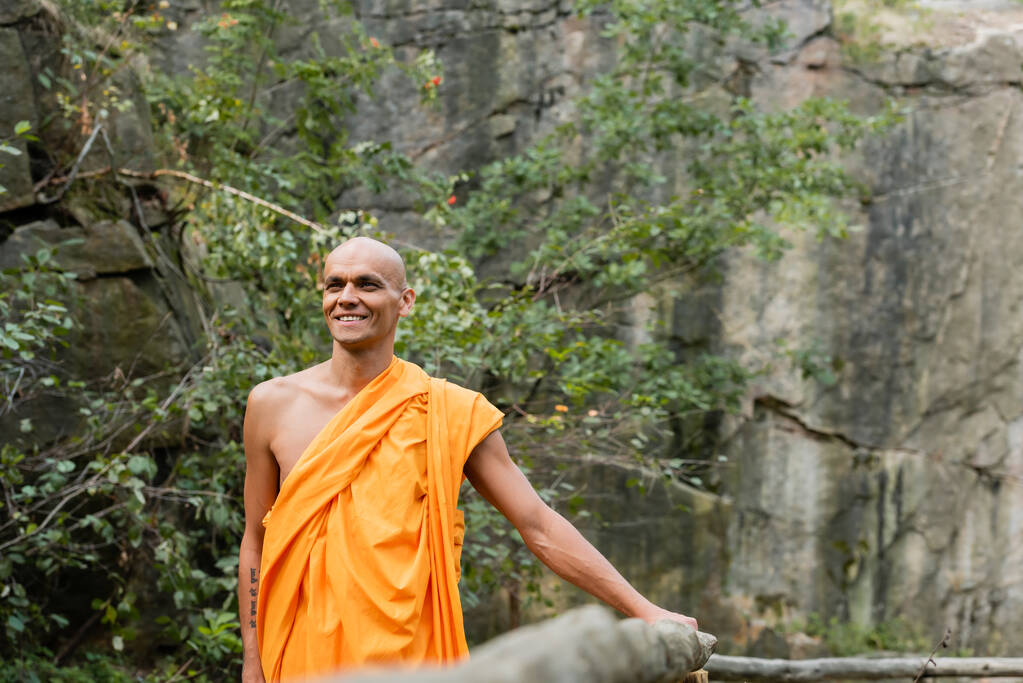 happy buddhist in orange kasaya walking near wooden fence in forest - Photo, Image