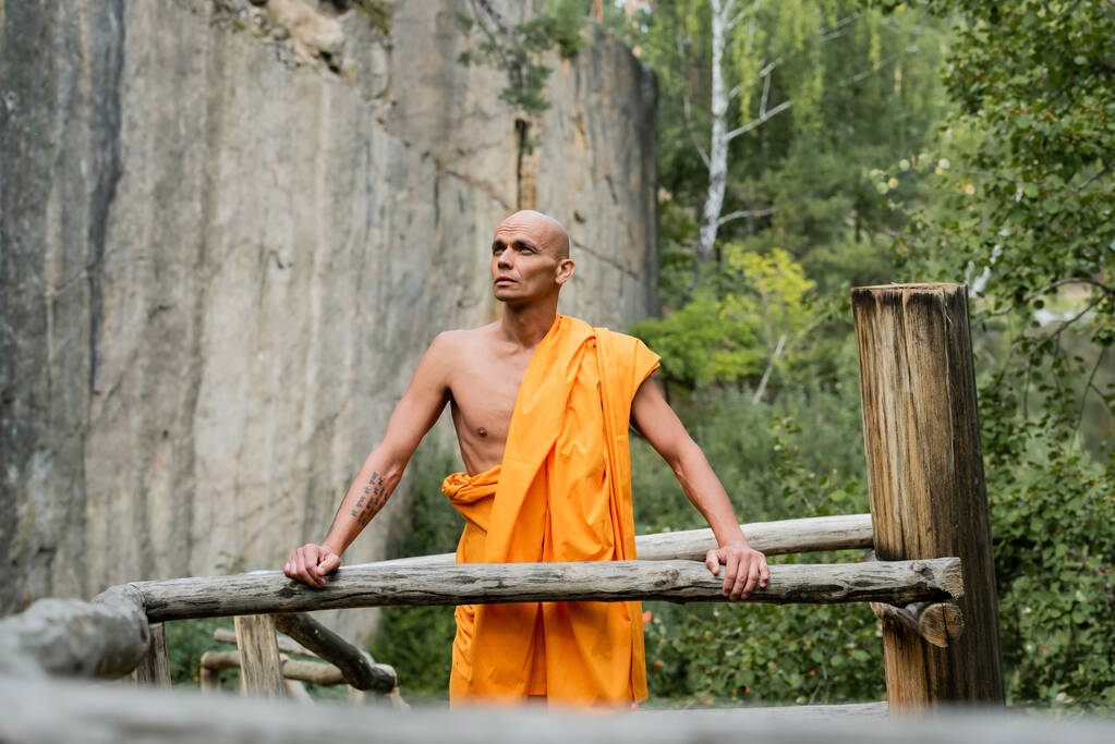 森の中の木の柵や岩のそばに見える伝統的な仏教の衣を着た男 - 写真・画像