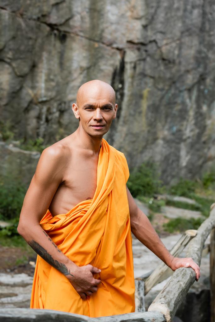 буддист в помаранчевій касі дивиться на камеру, стоячи біля дерев'яного паркану на відкритому повітрі
 - Фото, зображення
