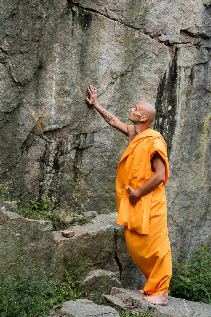 Buddhistischer Mönch in traditioneller orangefarbener Robe berührt Felsen - Foto, Bild