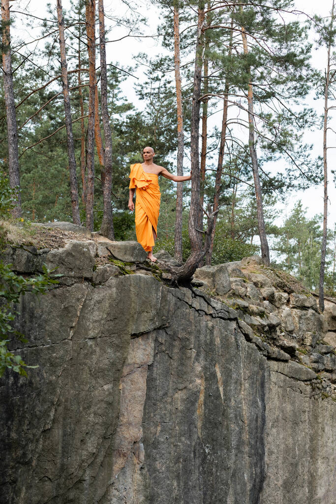 volledige weergave van boeddhistische monnik in oranje kasaya wandelen op rotsachtige klif in het bos - Foto, afbeelding