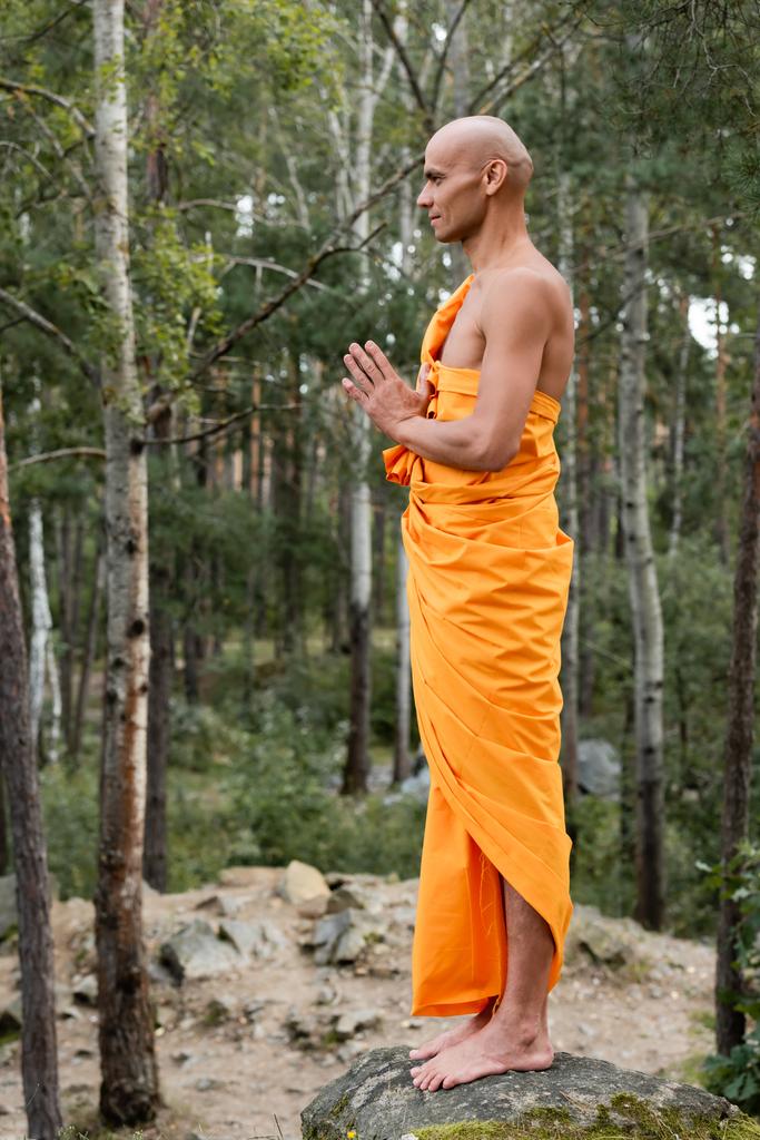 vue latérale du bouddhiste pieds nus en kasaya orange priant en forêt - Photo, image