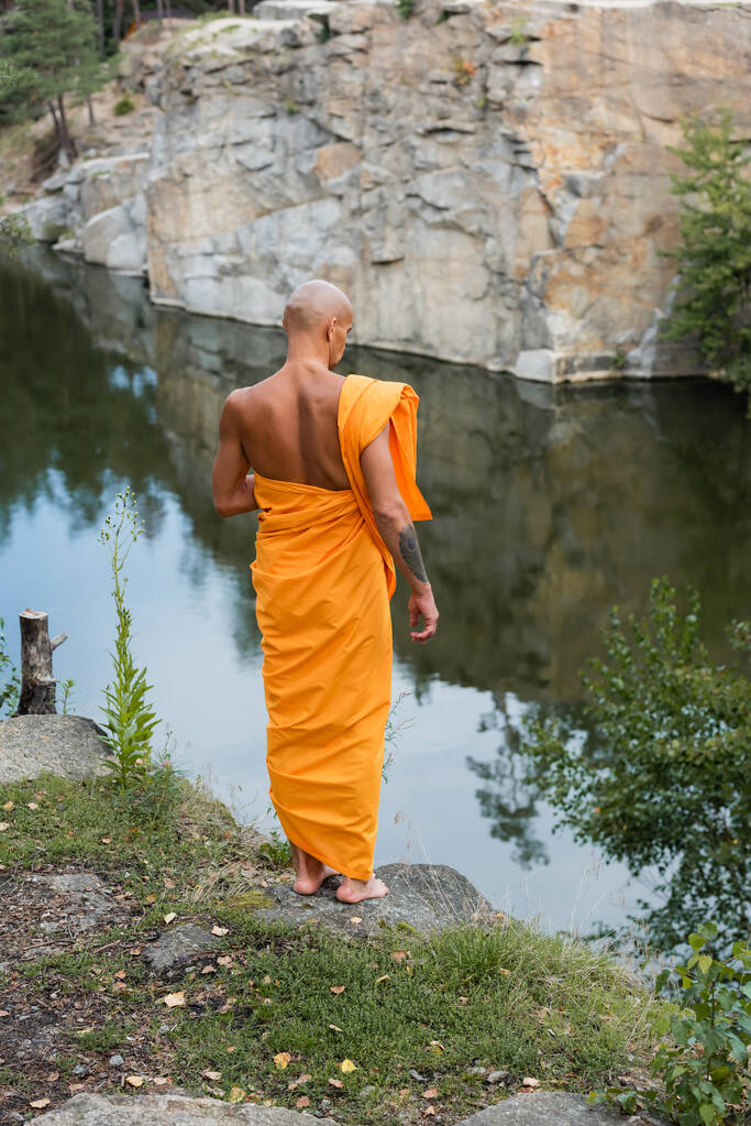 Rückansicht des Barfuß-Buddhisten in orangefarbenem Gewand, der am See im Wald meditiert - Foto, Bild
