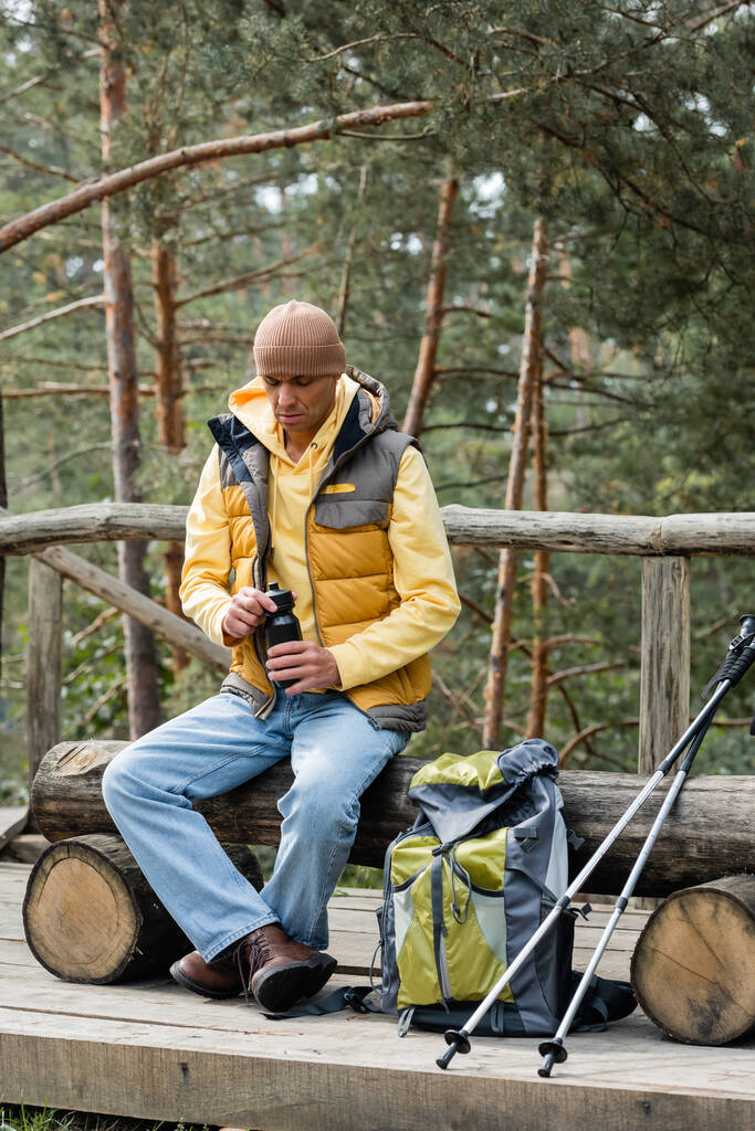 toerist in warm vest het openen van thermo fles tijdens het stoppen op houten bank in de buurt van rugzak en trekking palen - Foto, afbeelding