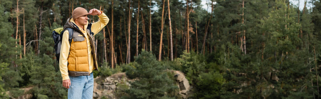 wędrowiec w ciepłej kamizelce i czapce trzymający się za rękę przy czole i odwracający wzrok w lesie, baner - Zdjęcie, obraz