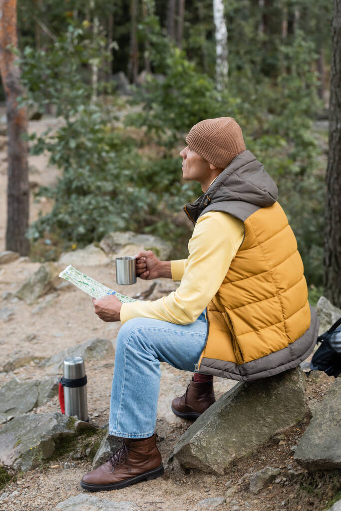 hiker in warm vest and beanie looking away while sitting with mug and map in forest - Photo, Image