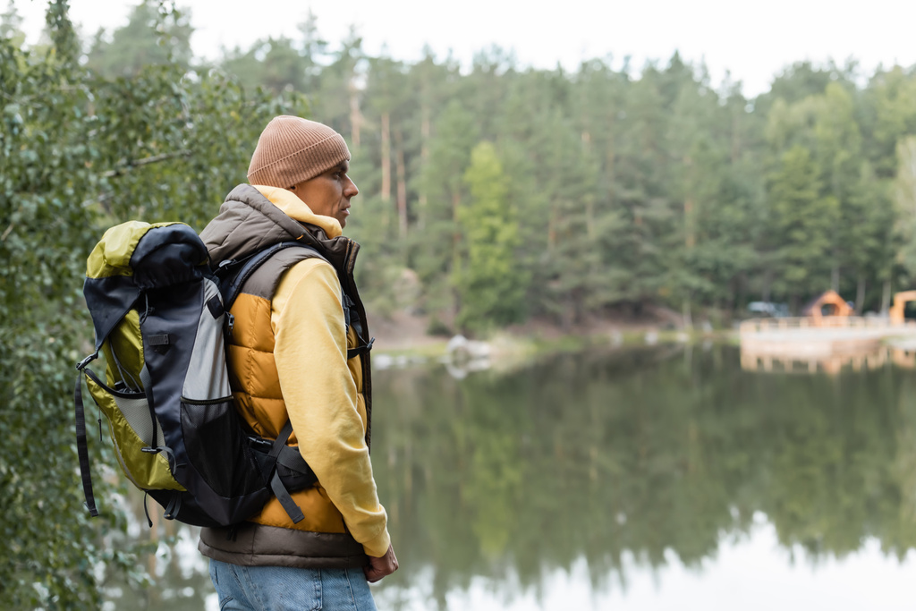 hiker with backpack looking away while standing near lake in forest - Photo, Image
