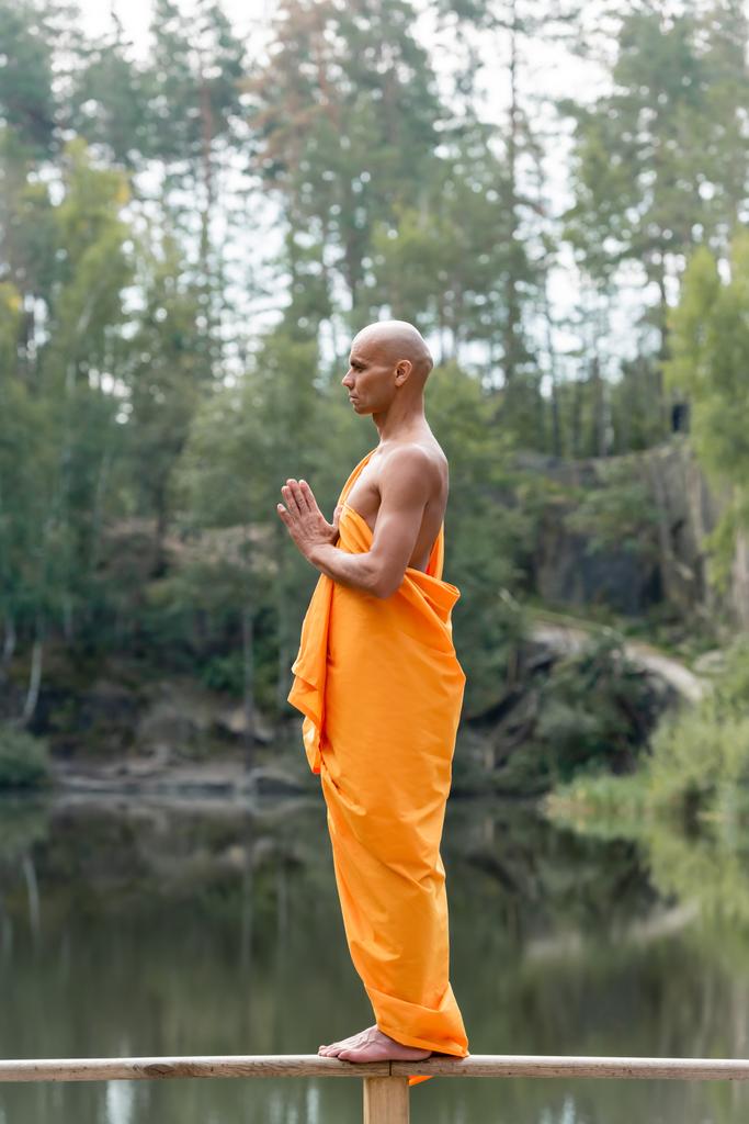 barefoot buddhist standing on wooden fence in forest while meditating with praying hands - Photo, Image