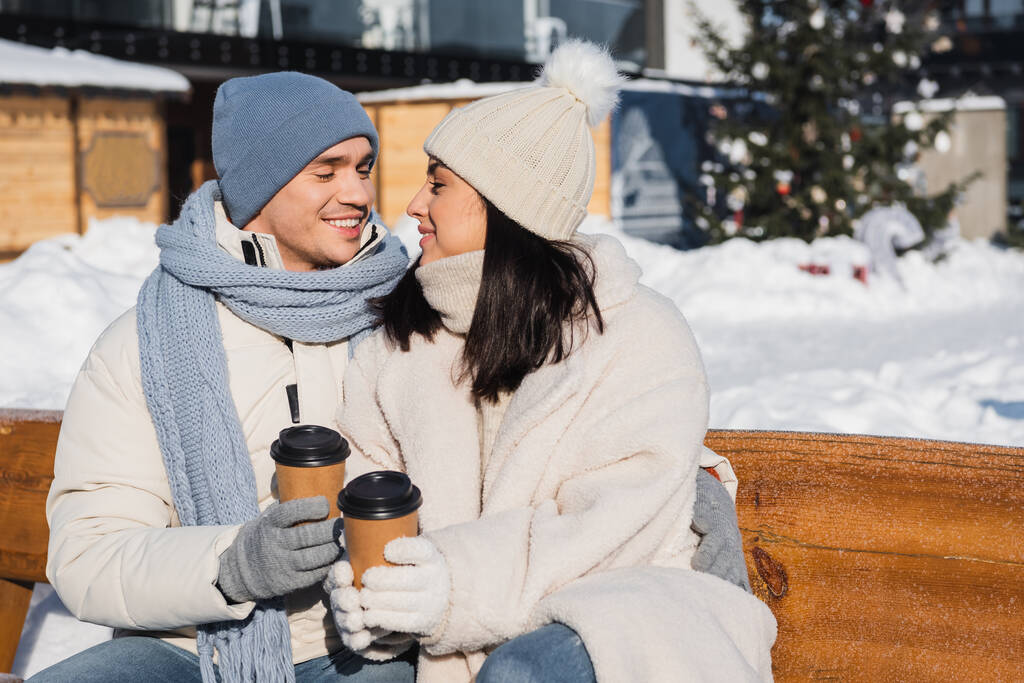 heureux homme et femme en bonnets tricotés se regardant tout en tenant des tasses en papier  - Photo, image