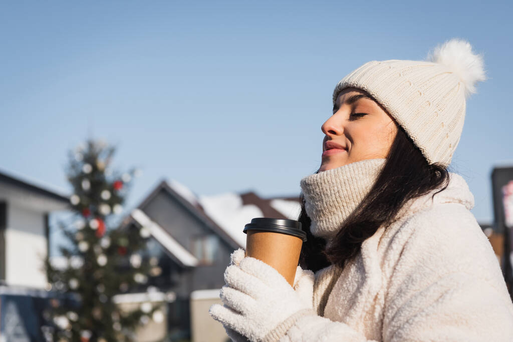 donna allegra con gli occhi chiusi e cappello invernale che tiene tazza di carta all'esterno  - Foto, immagini