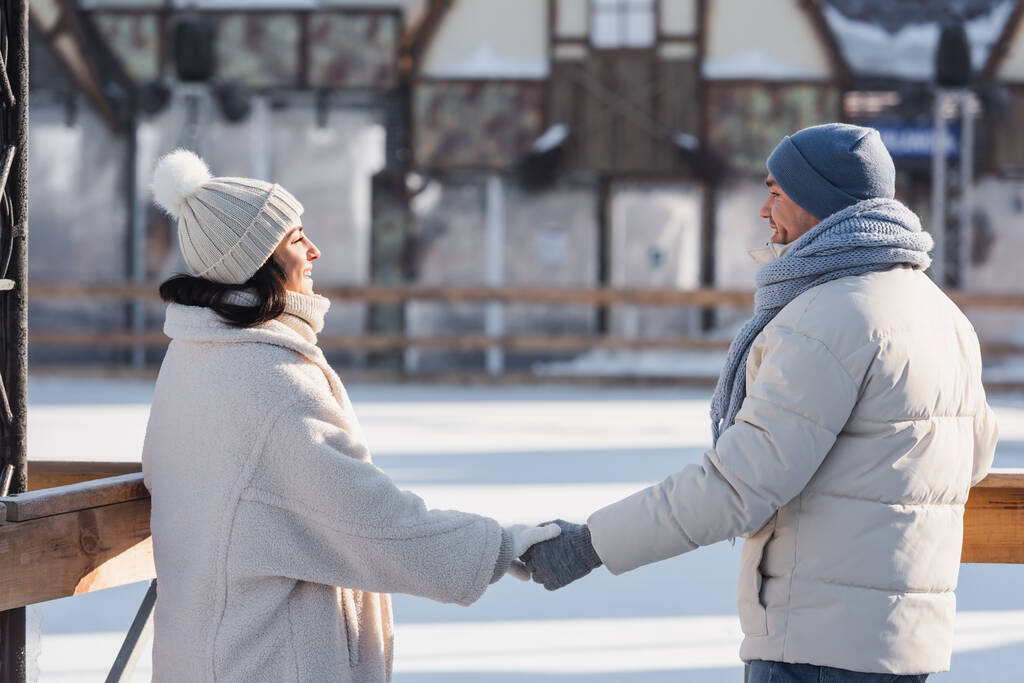 glückliches junges Paar mit Wintermützen, die einander anschauen, während sie sich draußen an den Händen halten - Foto, Bild