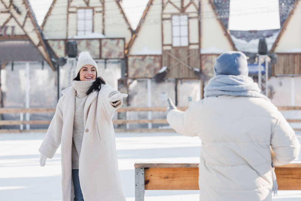 χαμογελαστή νεαρή γυναίκα σε χειμερινό καπέλο στέκεται με απλωμένο χέρι κοντά θολή φίλο  - Φωτογραφία, εικόνα