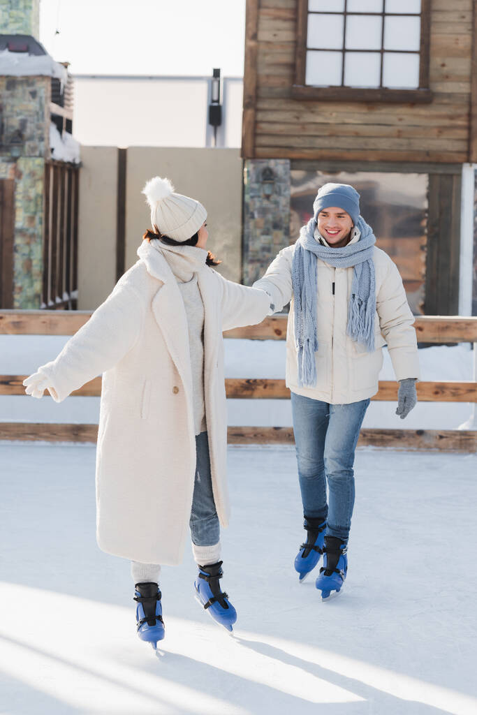 longitud completa de feliz joven en sombrero de invierno y patines de hielo de la mano con su novia en pista de hielo - Foto, Imagen