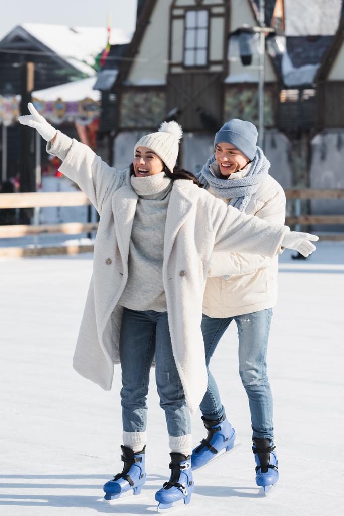 longitud completa de hombre joven feliz en sombrero de invierno detrás de la novia sonriente con las manos extendidas en pista de hielo - Foto, imagen